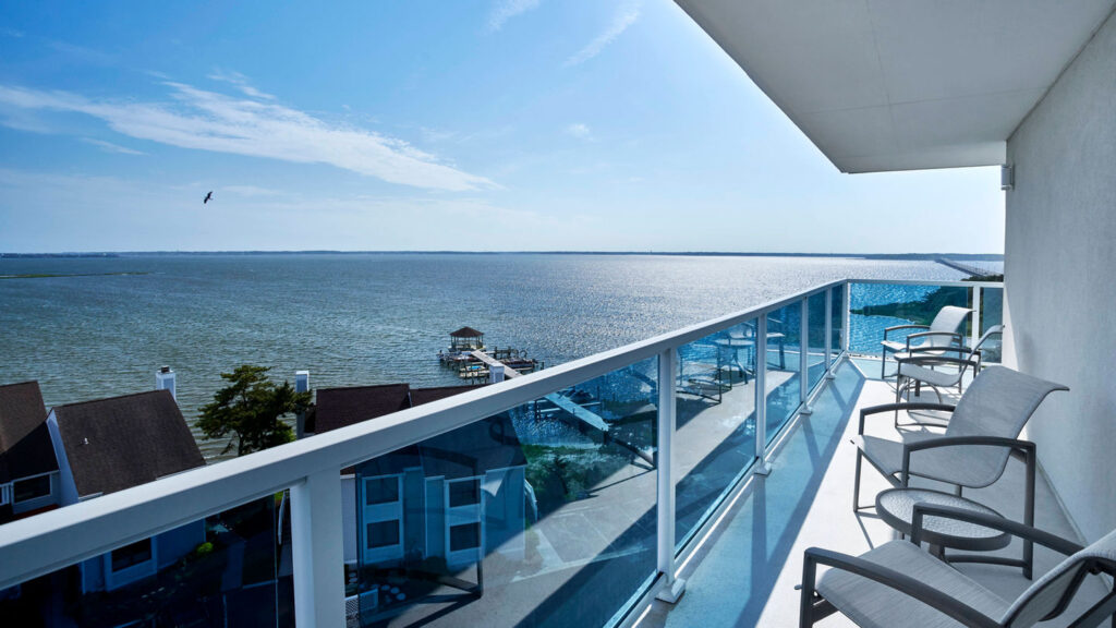 A private balcony with glass railings and lounge chairs overlooking the calm waters of the Chesapeake Bay on a sunny day, with a dock and waterfront homes visible below.