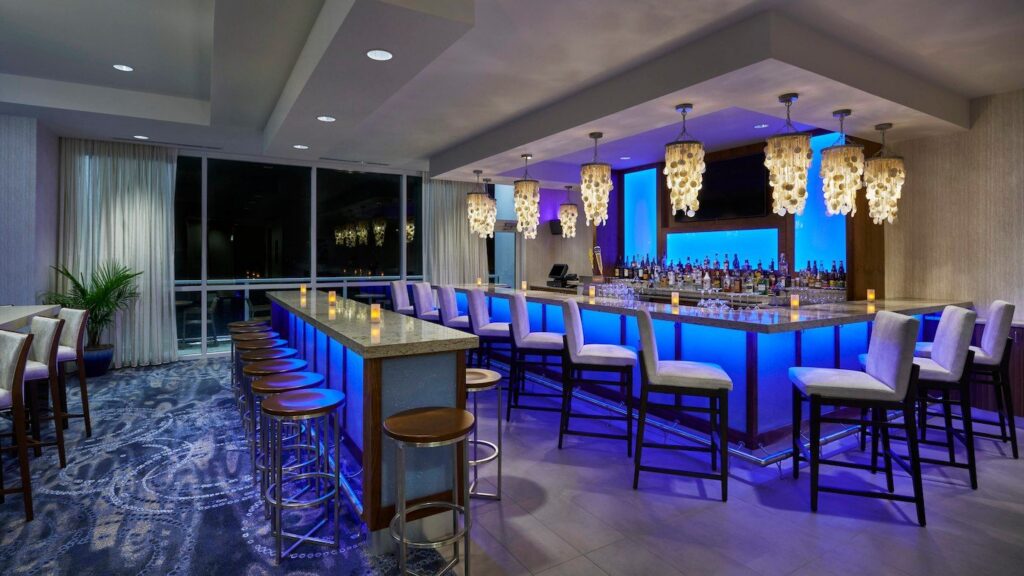 An upscale hotel bar with a U-shaped marble counter, backlit blue base, shell chandelier pendant lights, and floor-to-ceiling windows overlooking the water at night.
