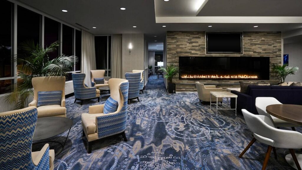 A spacious hotel lounge with patterned blue carpeting, mixed seating arrangements, tropical plants, and a wide linear gas fireplace set into a stone accent wall.