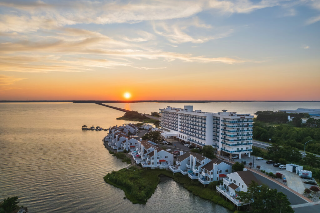 Aerial drone view of the Residence Inn Ocean City at golden hour, situated on a narrow peninsula surrounded by the bay, with a vivid orange and yellow sunset reflected on the water.