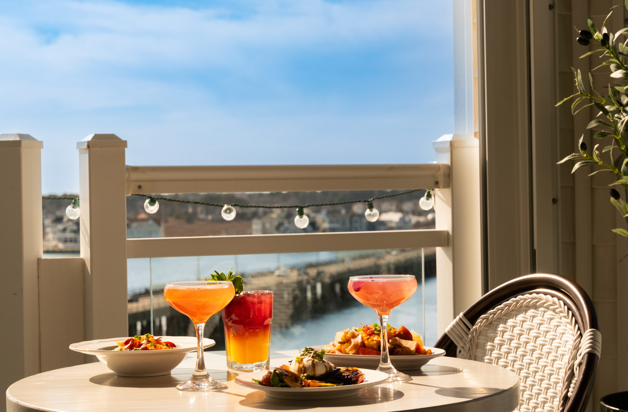 A sunny outdoor balcony table overlooking a harbor, featuring colorful cocktail drinks and plated tapas against a clear blue sky.