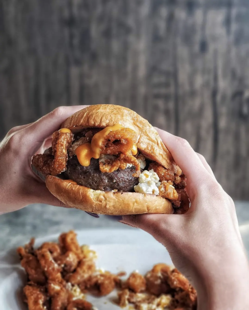 Two hands holding a thick burger on a soft bun, loaded with fried calamari rings, melted cheese, and a drizzled creamy sauce, set against a dark rustic wood backdrop with a side of fried appetizers blurred below.