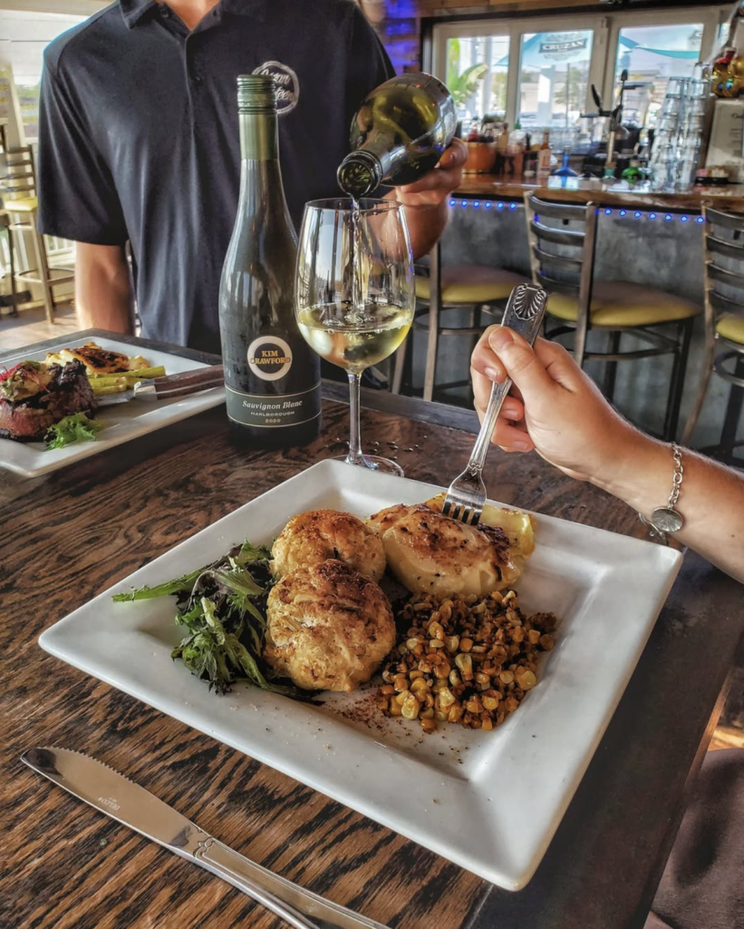 A dining table featuring a white square plate with two golden-brown crab cakes, roasted corn, a baked potato, and a side salad. A diner holds a fork over the plate while a server pours white wine into a glass next to a bottle of Kim Crawford Sauvignon Blanc.