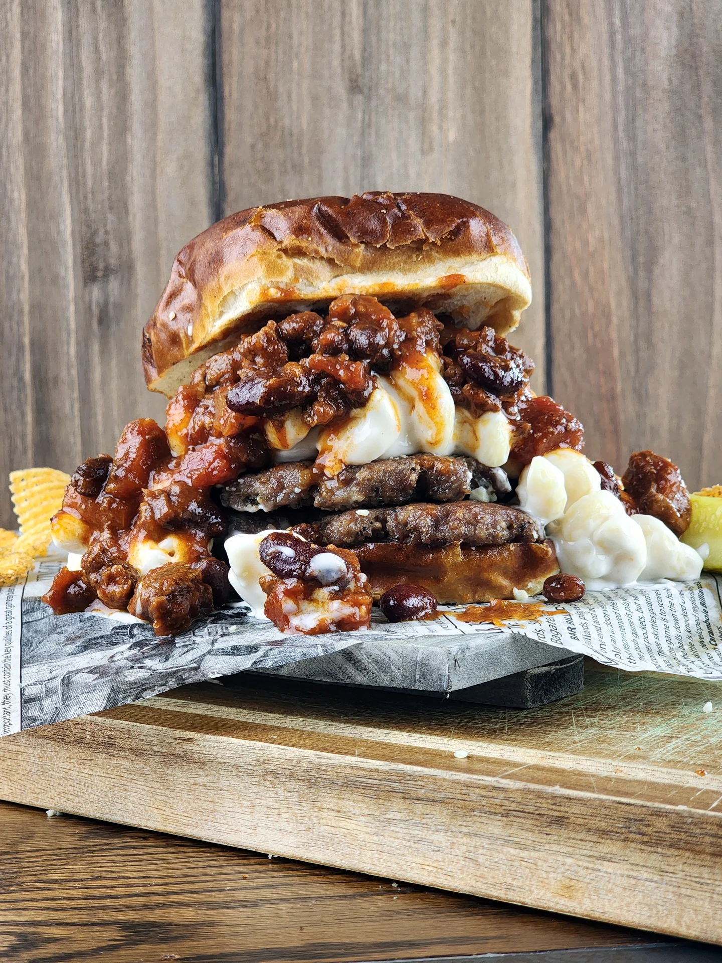 A stacked double beef burger on a toasted brioche bun, loaded with mac and cheese and topped with a chili sauce containing red kidney beans, served on newspaper-printed wax paper over a wooden board.