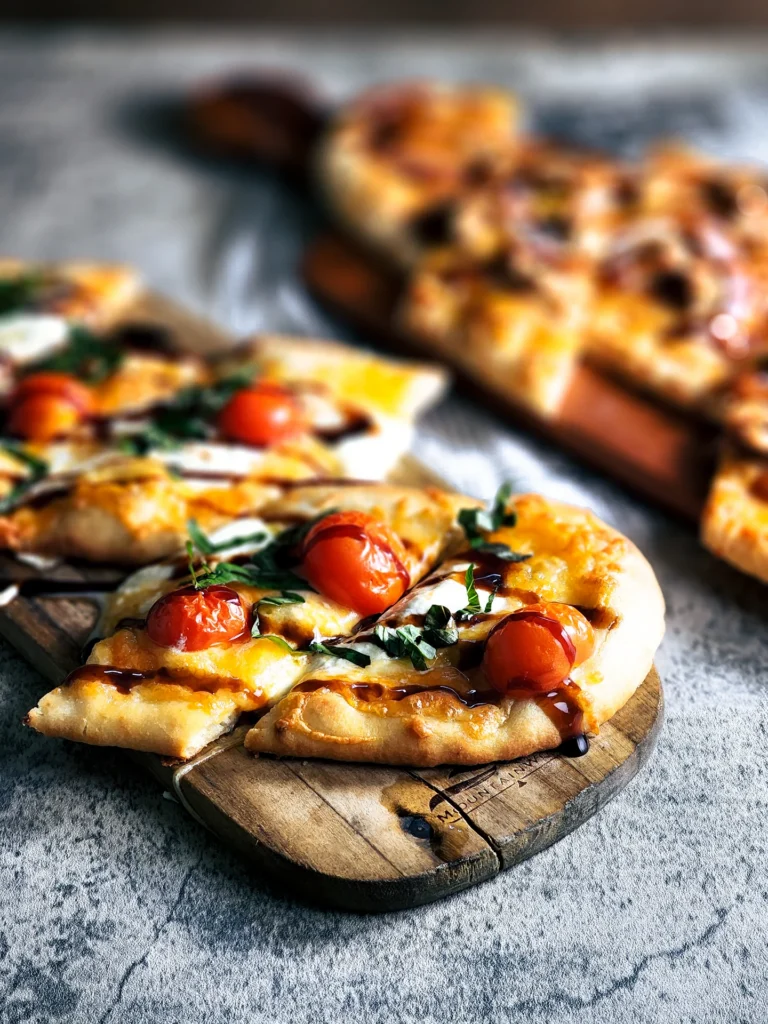 A flatbread pizza sliced on a wooden board, topped with melted cheese, fresh cherry tomatoes, basil, and drizzled with a dark balsamic reduction.