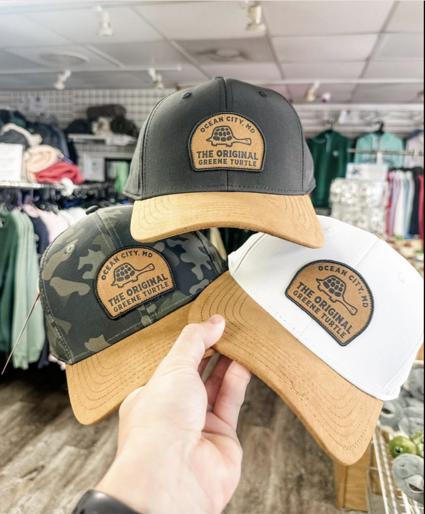 A hand holding three baseball caps fan-style in front of a retail store background. The hats come in black, camouflage, and white, each featuring a tan suede brim and a matching leather patch with a turtle graphic that reads "Ocean City, MD - The Original Greene Turtle."
