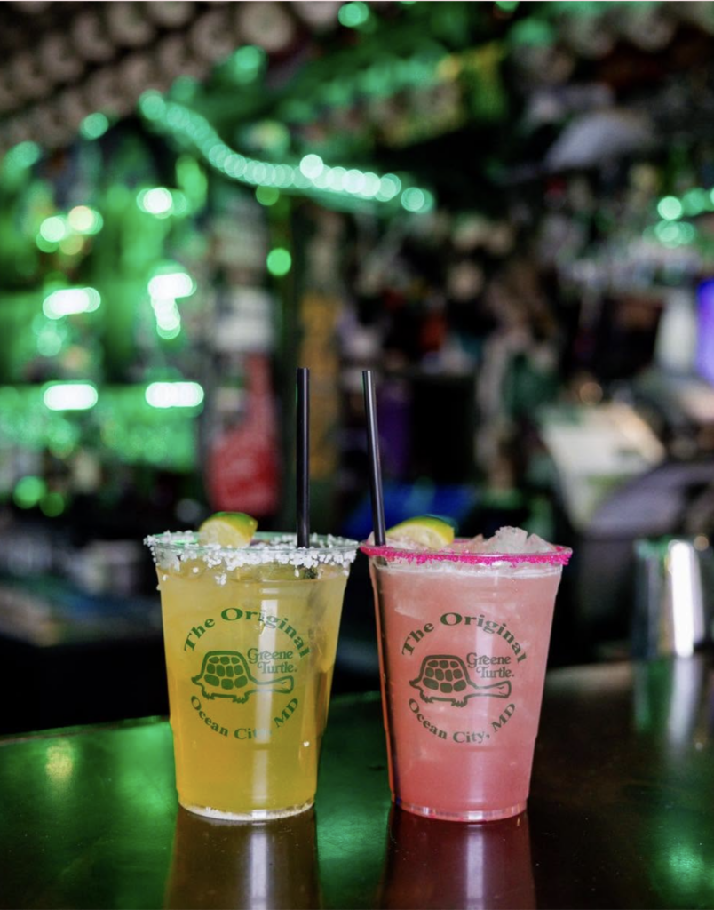Two clear plastic cocktail cups featuring "The Original Greene Turtle Ocean City, MD" logo sitting on a bar. The drink on the left is yellow with a salted rim and lime slice, while the drink on the right is pink with a red sugar rim and lime slice. Both have black straws, set against a background illuminated by green bar lights.