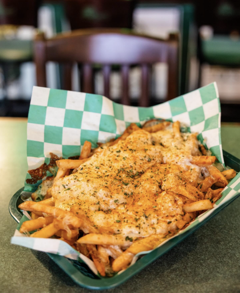 A green basket lined with green and white checkered paper filled with French fries topped with a generous layer of melted cheese, seasoning, and herbs. A wooden bar chair is visible blurred in the background.