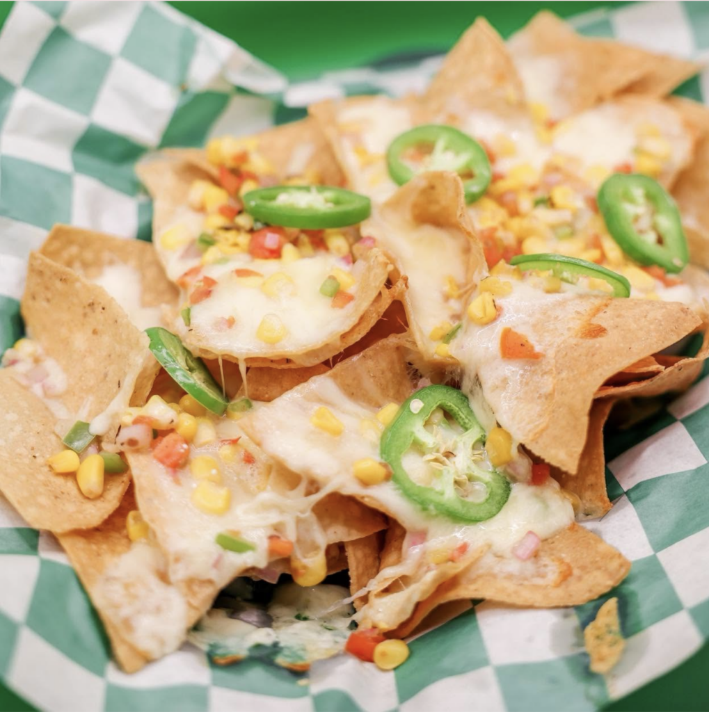 A close-up shot of tortilla chip nachos served in a green-and-white checkered basket, layered with melted white cheese, diced red and yellow peppers, corn, and topped with fresh sliced jalapeños.