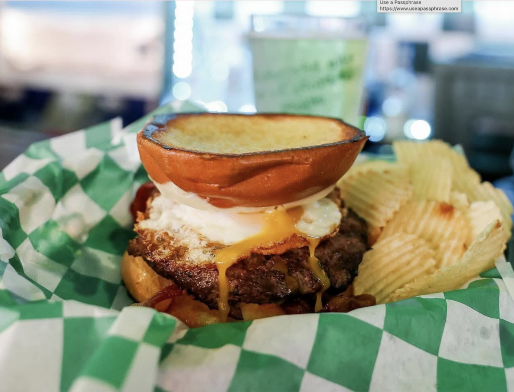 A juicy burger with a fried egg and melted cheese running down the patty, served on a toasted bun in a green-and-white checkered basket with a side of ridged potato chips.