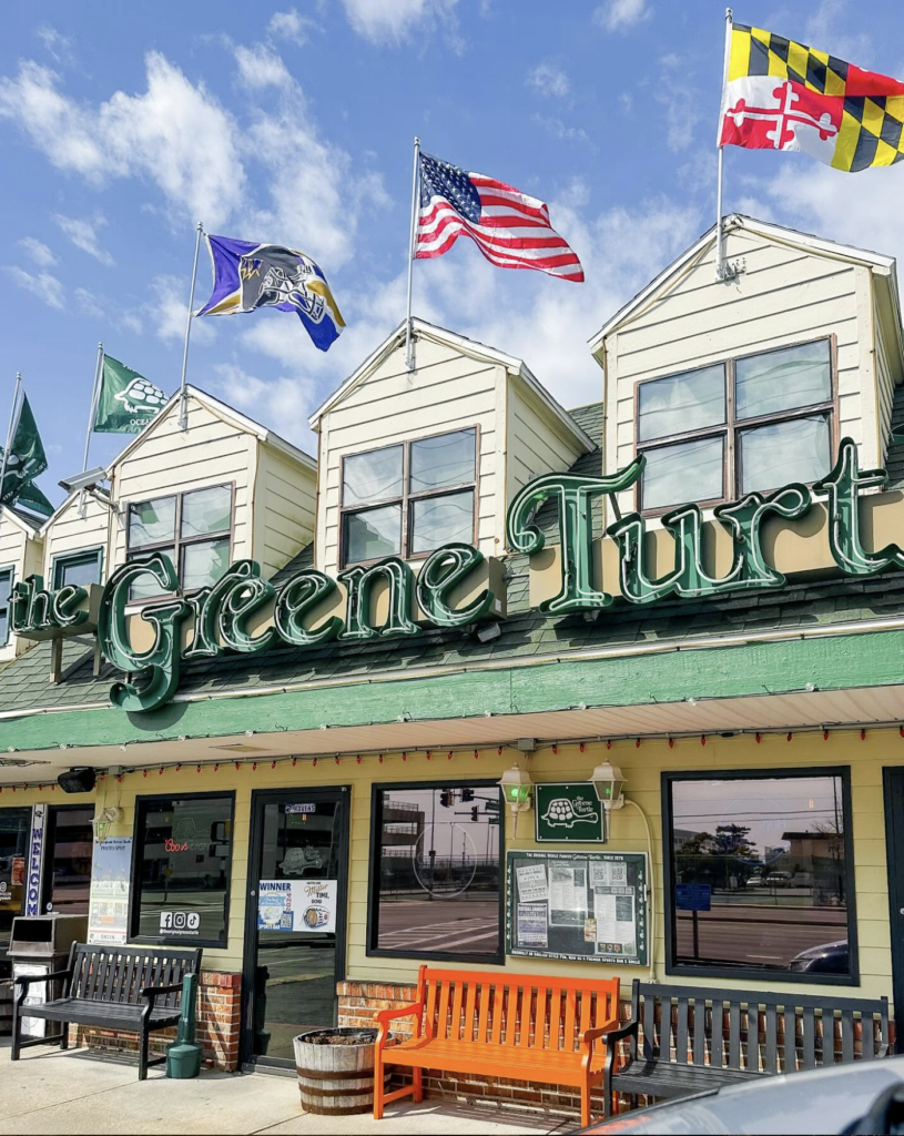 Exterior view of The Original Greene Turtle restaurant building featuring green neon signage, several flags flying along the roofline (including the US, Maryland, and sports flags), and orange and black wooden benches parked along the sidewalk out front.